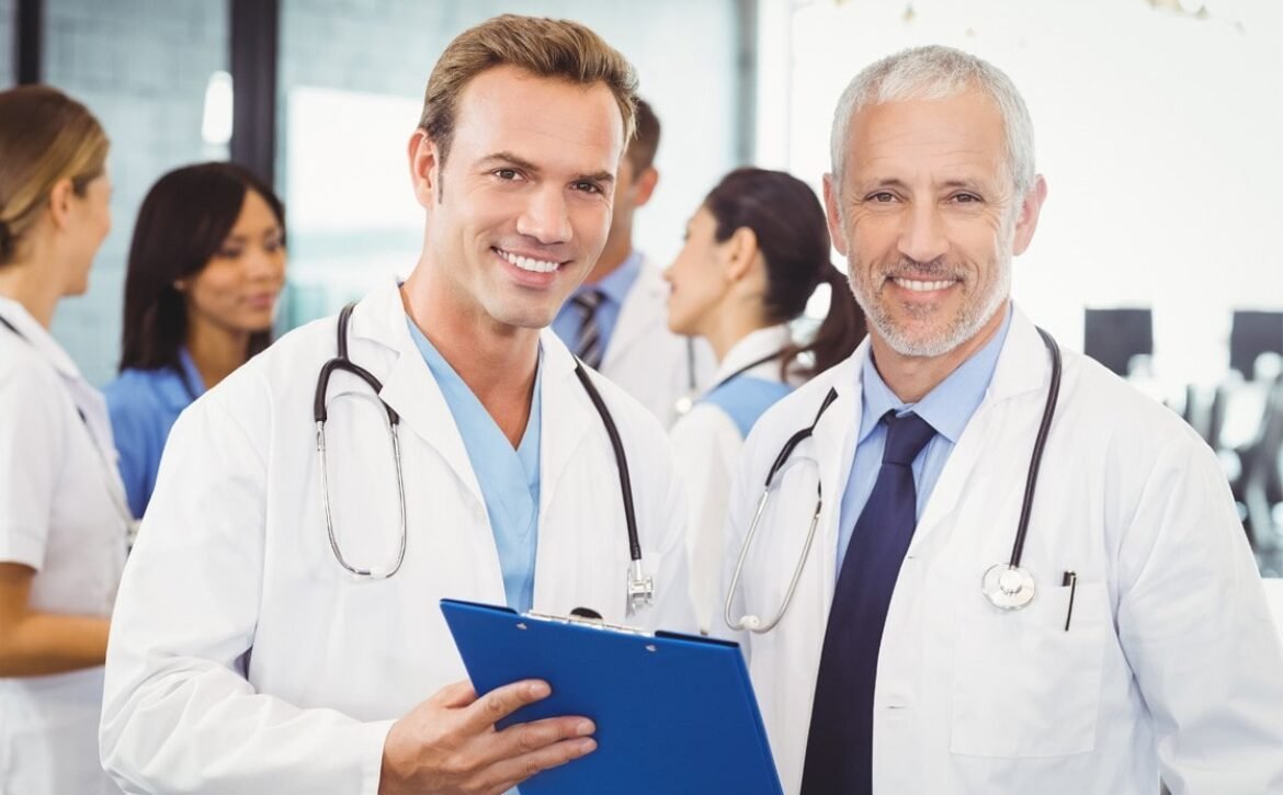 Two male doctors, smiling and holding a clipboard, stand together in a medical office with other healthcare professionals in the background—perfect teammates for MCCQE1 exam prep with Ace QBank.