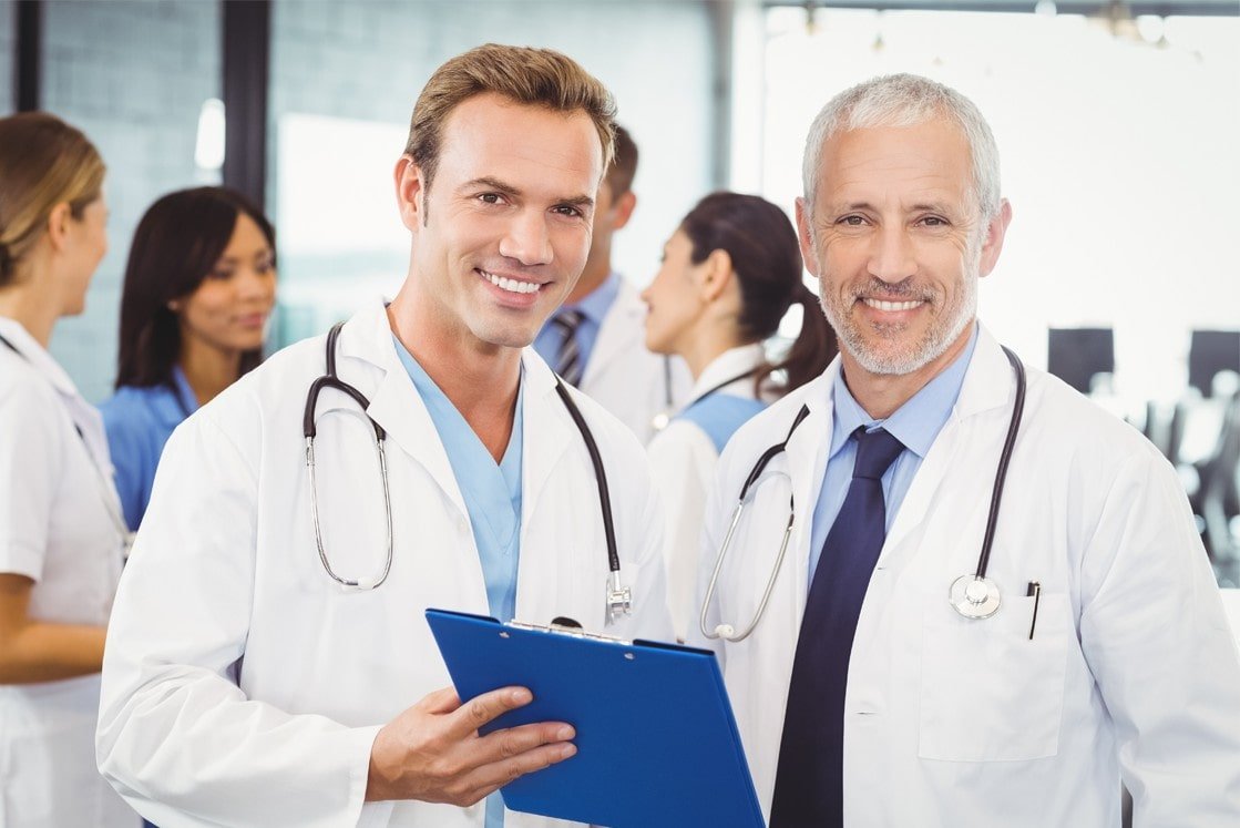 Two male doctors, smiling and holding a clipboard, stand together in a medical office with other healthcare professionals in the background—perfect teammates for MCCQE1 exam prep with Ace QBank.