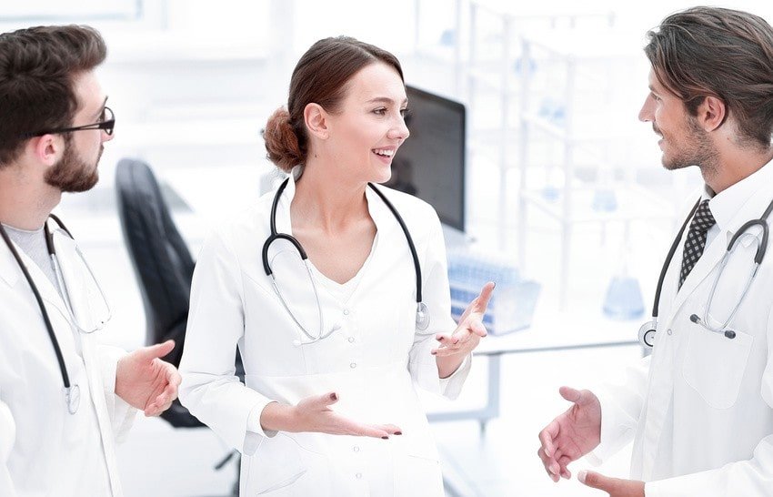 Three doctors in white coats with stethoscopes around their necks are having a discussion in a bright medical office, sharing tips on MCCQE1 exam prep and the benefits of using Ace QBank for their studies.