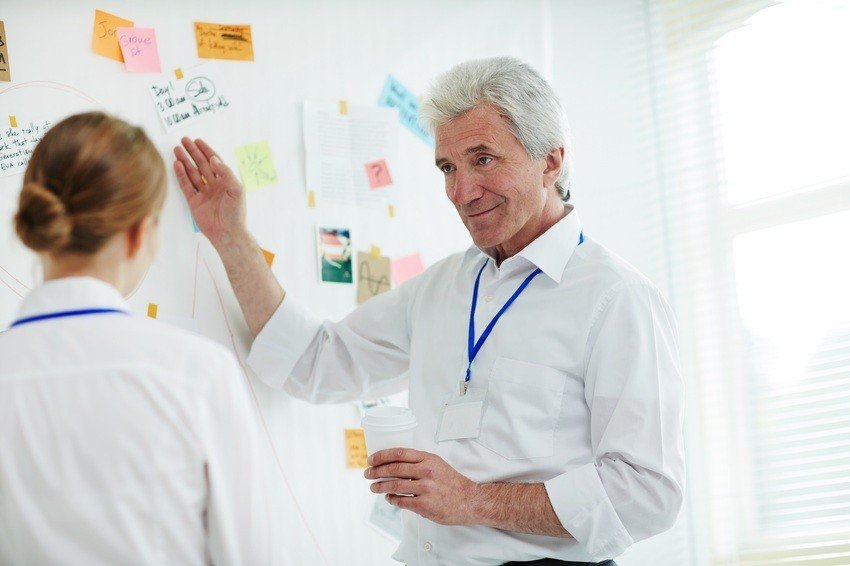 An older man in a white shirt gestures toward a whiteboard covered with notes and sticky notes, discussing Ace QBank's MCCQE1 exam prep strategies with a colleague.