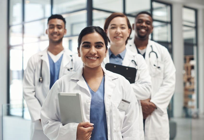 Four medical professionals in lab coats stand together indoors, smiling and holding clipboards, with a modern hospital or clinic setting in the background, to get their medical licence in Canada.