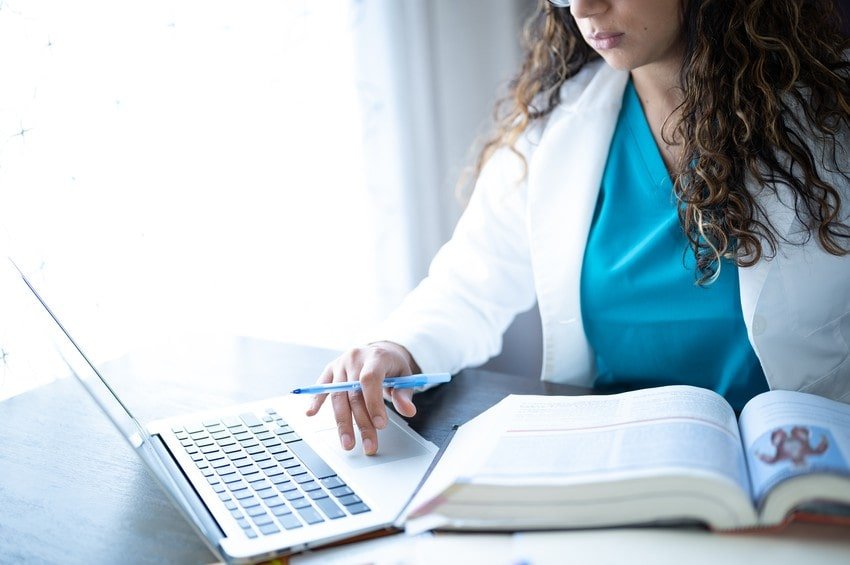 A person in medical scrubs and a white coat uses a laptop and takes notes from an open book at a desk, focused on MCCQE1 Exam Prep with Ace QBank.