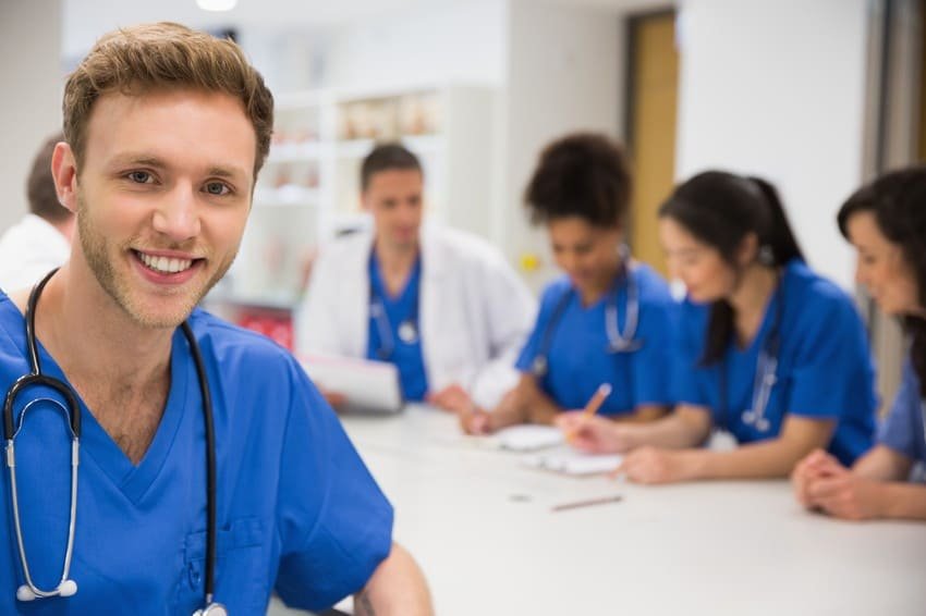 A smiling healthcare worker in blue scrubs stands in the foreground, while four colleagues discuss MCCQE Part 1 Prep and Ace QBank strategies for the MCCQE1 exam at a table in the background.
