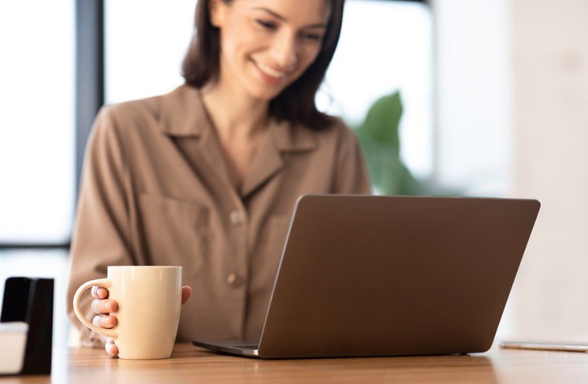 A woman smiles while using a laptop at a desk, holding a white coffee mug in her left hand, as she reviews MCCQE1 exam prep materials from the Ace QBank Canadian question bank.