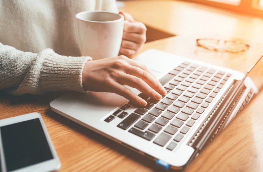 A person types on a laptop keyboard with one hand while holding a white mug in the other, next to a smartphone, Ace QBank notes, and a pair of glasses on a wooden desk. This Canadian question bank is perfect for MCCQE1 exam prep.