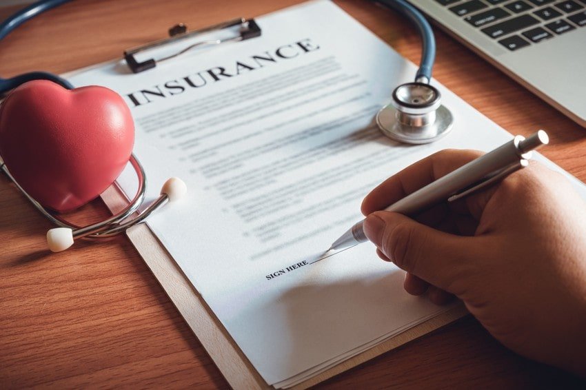 A hand signs an insurance document on a clipboard, with a stethoscope, a heart model, and a laptop on the wooden desk. Perfect for those who passed the MCCQE1 exam with Ace QBank and are learning about doctor salaries.