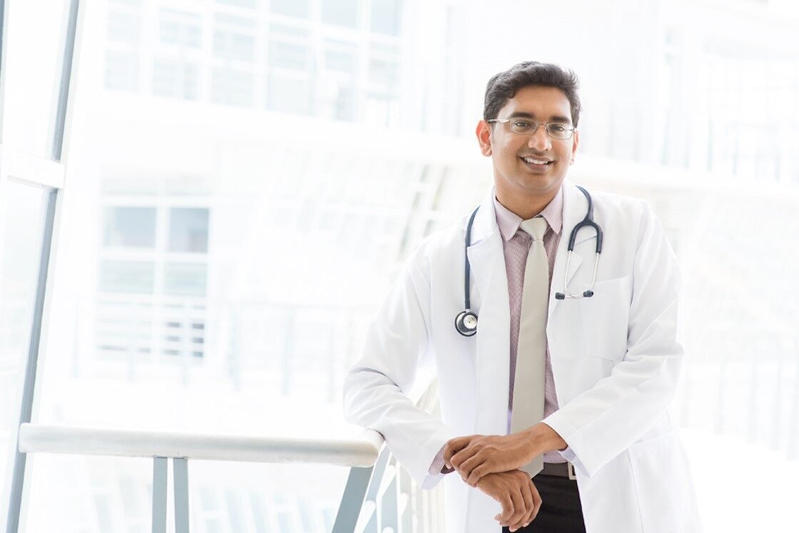 A doctor wearing a white coat and stethoscope stands indoors, leaning on a railing and smiling at the camera, confident after using Ace QBank for MCCQE1 Exam Prep.
