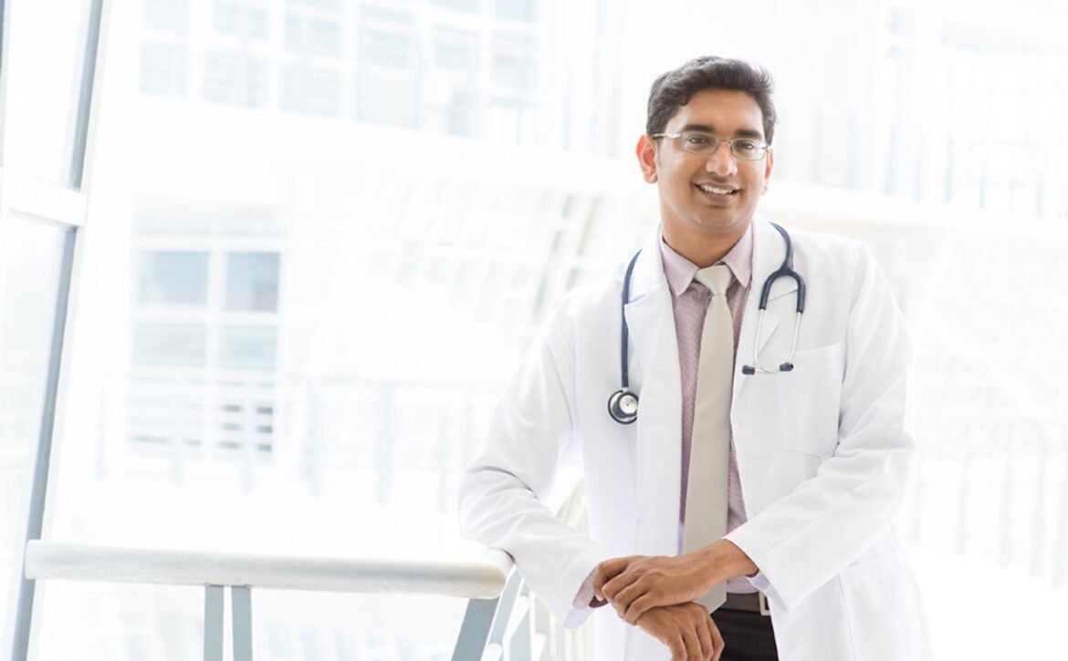 A doctor wearing a white coat and stethoscope stands indoors, leaning on a railing and smiling at the camera, confident after using Ace QBank for MCCQE1 Exam Prep.