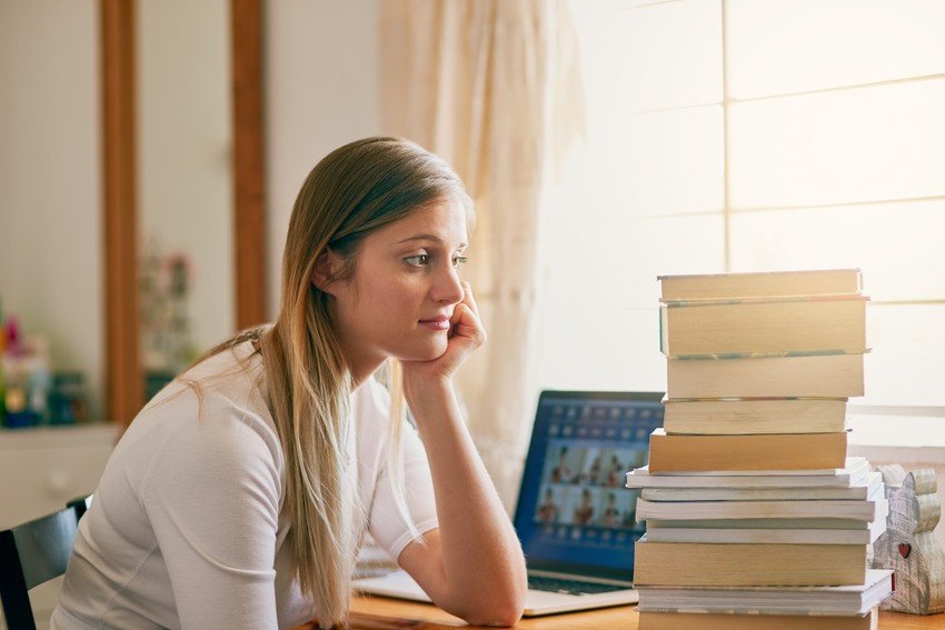 A woman sits at a desk with her chin resting on her hand, looking at a stack of books next to a laptop with an Ace QBank website open for MCCQE1 exam prep on the screen.