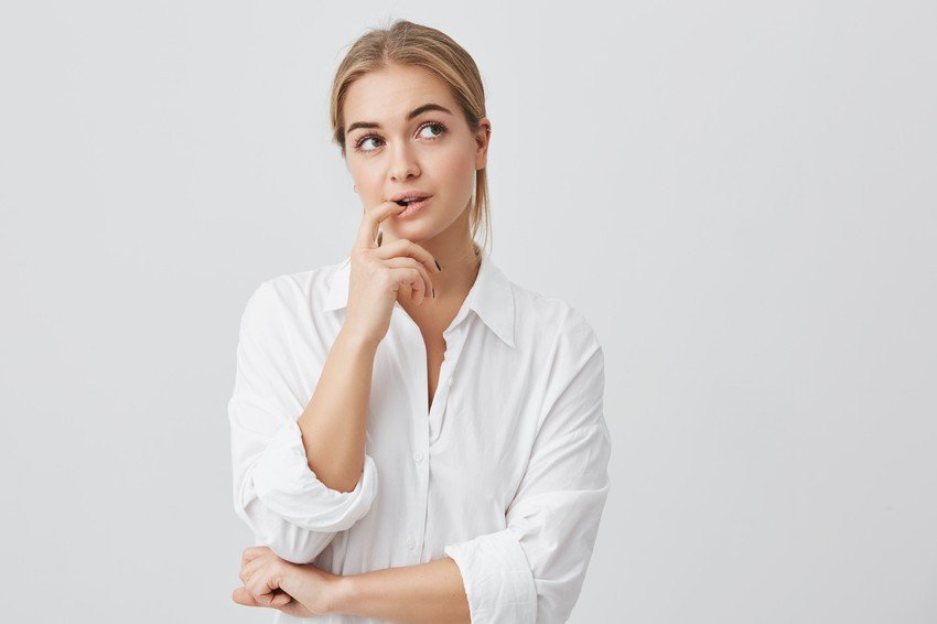 A woman in a white shirt stands against a plain background, looking to the side with her finger on her lips, appearing thoughtful. Much like someone deep in MCCQE1 exam prep or exploring a Canadian question bank.