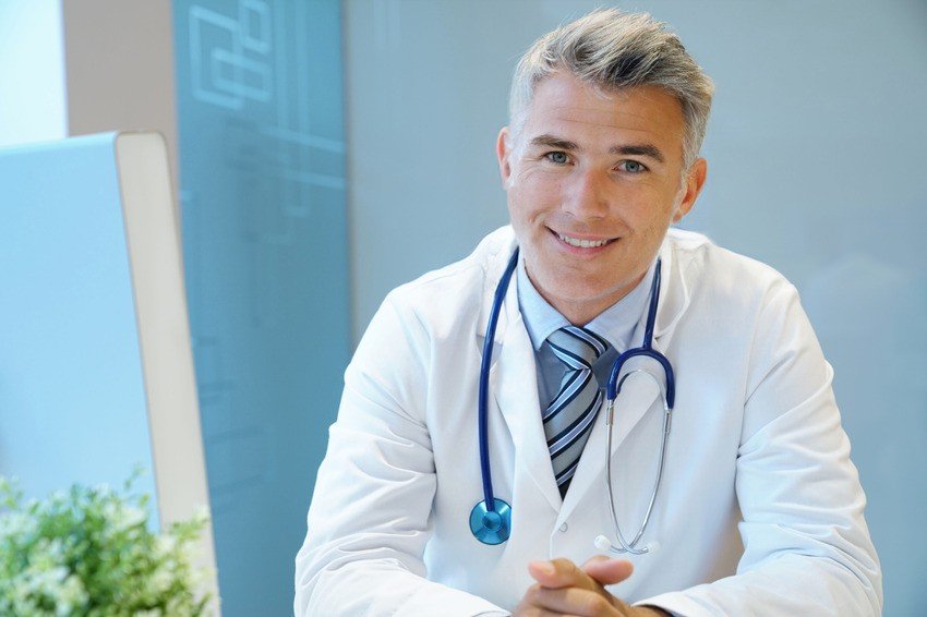 A male doctor with gray hair, wearing a white coat and stethoscope, sits at a desk and smiles at the camera, possibly offering insights on MCCQE1 exam prep and Ace QBank.