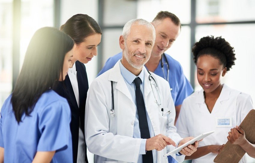 A group of healthcare professionals, including doctors and nurses, stand together in a hospital setting, reviewing Medicine in Canada resources on a digital tablet as part of their MCCQE1 exam Prep.