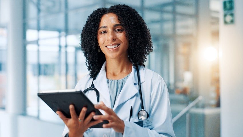 A doctor in Canada in a white coat with a stethoscope around her neck smiles while holding a digital tablet in a modern medical facility.