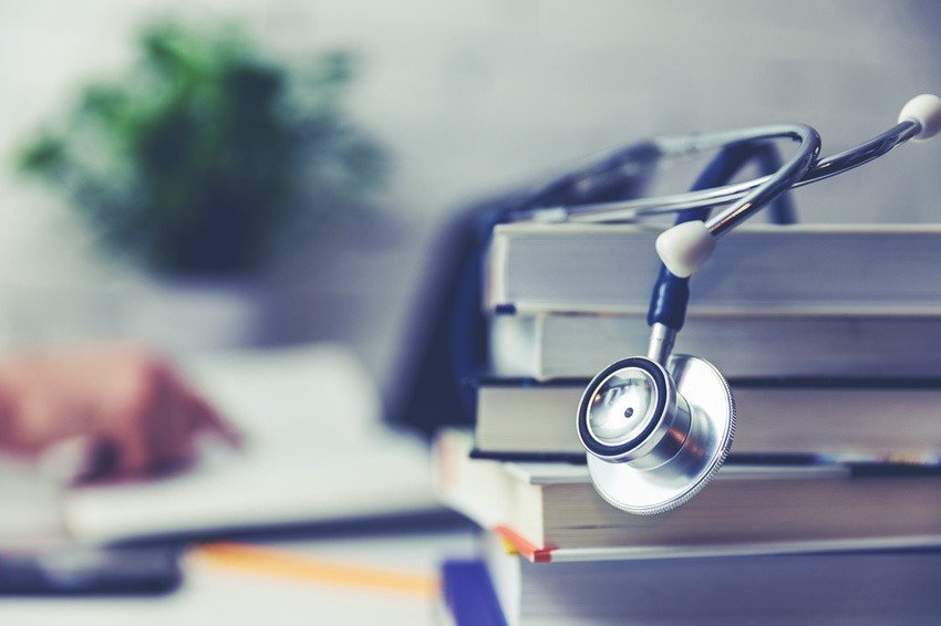 A stethoscope rests on a stack of books, suggesting the journey of a doctor in Canada, with blurred handwriting in the background and a green plant in soft focus.