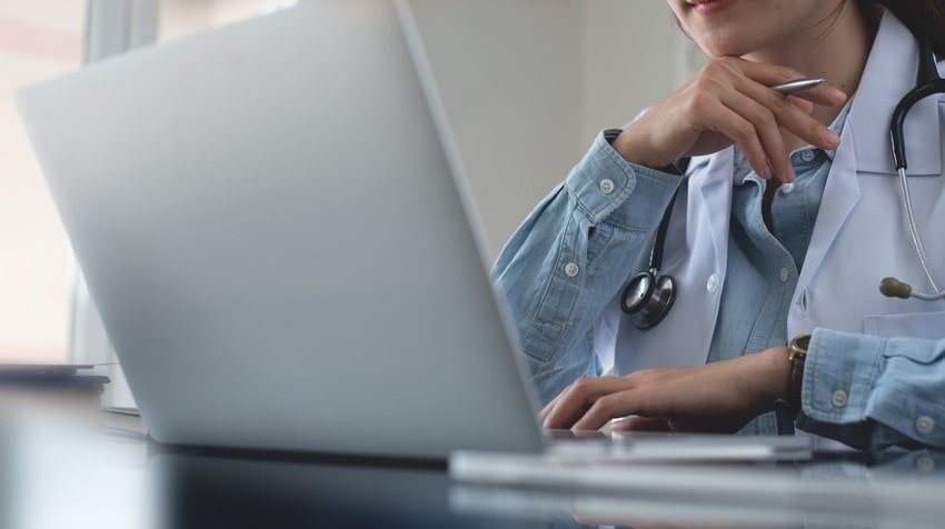 A Doctor in Canada with a stethoscope sits at a desk, working on a laptop computer, possibly reviewing practice questions from Ace QBank.