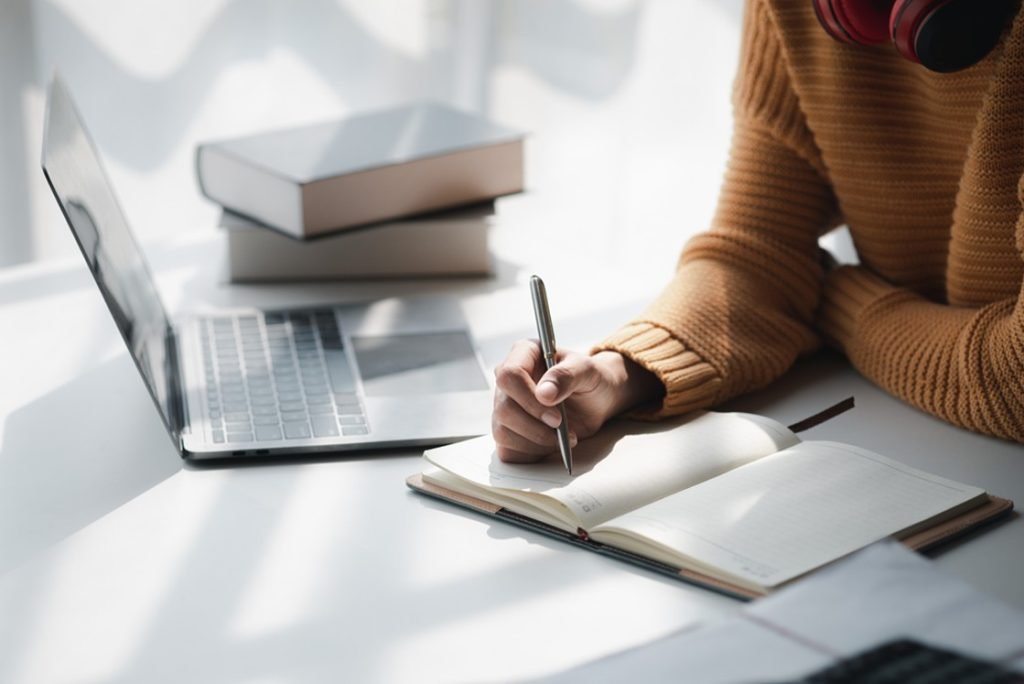 Person in a brown sweater writes in a notebook at a desk with an open laptop and stacked books nearby, focusing on MCCQE1 Exam Prep using the Ace QBank.
