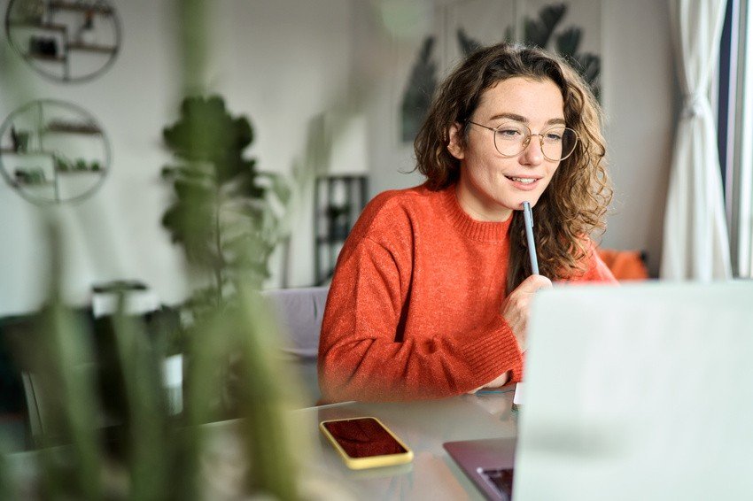 A woman wearing glasses and an orange sweater sits at a table with a laptop, reviewing MCCQE1 Exam Dates on the screen, a pen to her mouth and her phone beside her. Preparing efficiently with Ace QBank.