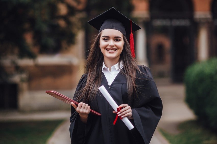 A young woman in a graduation cap and gown holds a diploma and a folder, smiling outdoors on a university campus, celebrating graduating college.
