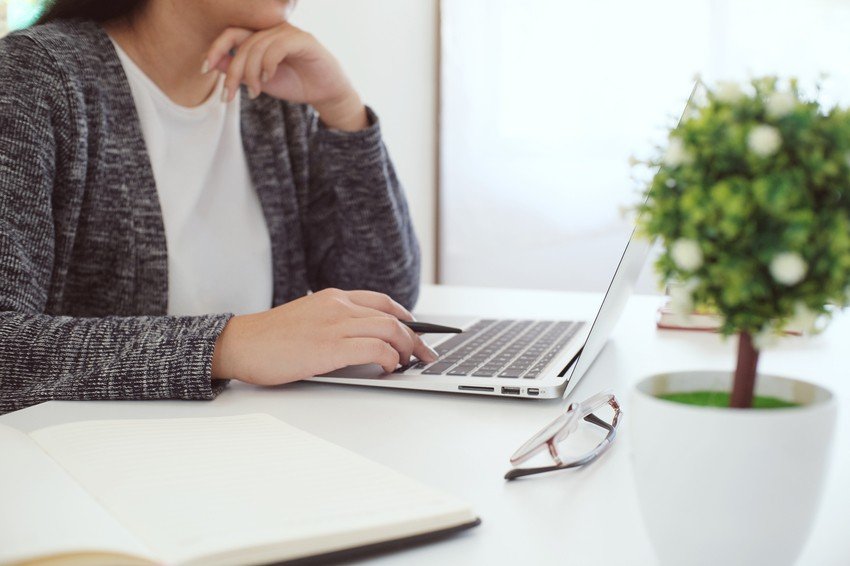 Person working at a desk with a laptop, notebook, eyeglasses, and a potted plant checks MCCQE1 Exam Dates on Ace QBank, resting their chin on one hand while typing with the other.