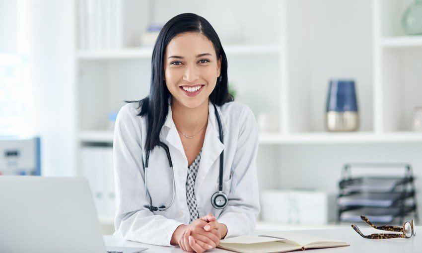 A doctor in a white coat sits at a desk with an open notebook, smiling at the camera. A stethoscope, laptop and eyeglasses are on the desk. Ready for focused MCCQE1 exam prep with Ace QBank.