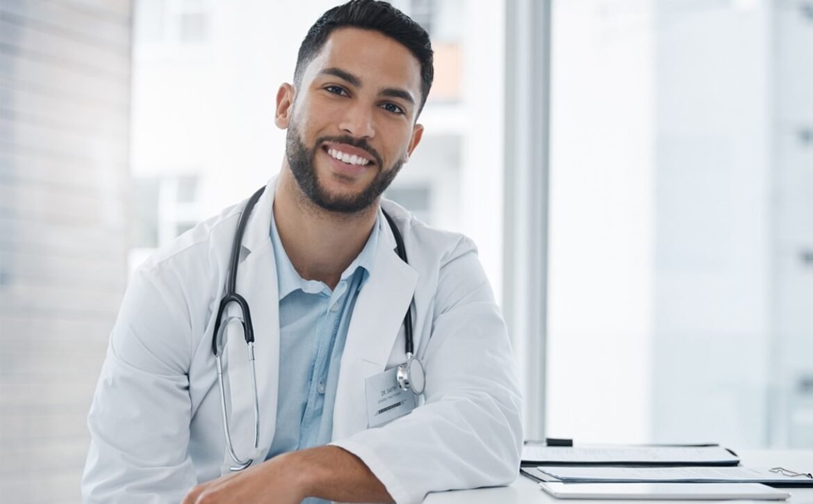 A male doctor wearing a white coat and stethoscope smiles while seated at a desk with an Ace QBank account open for the MCCQE1 exam prep and paperwork in a bright office.