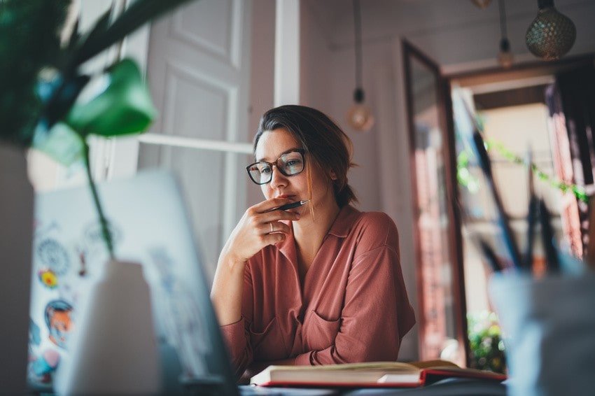 A woman wearing glasses sits at a desk, looking intently at her laptop, with an open notebook and pen in hand in a well-lit room, reviewing her MCCQE1 study plan on Ace QBank.