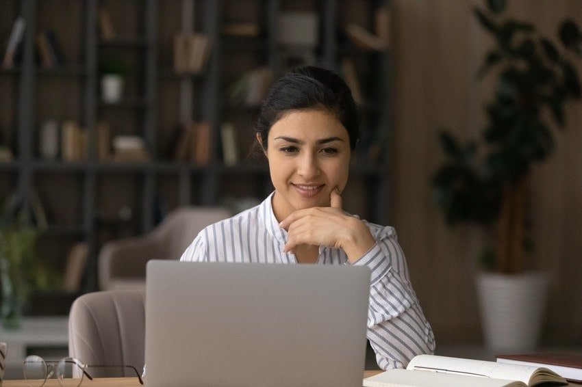 Woman sitting at a desk in front of a laptop, smiling with her hand on her chin. She is studying for MCCQE1 and wants to know how to use Ace QBank effectively for MCCQE1.