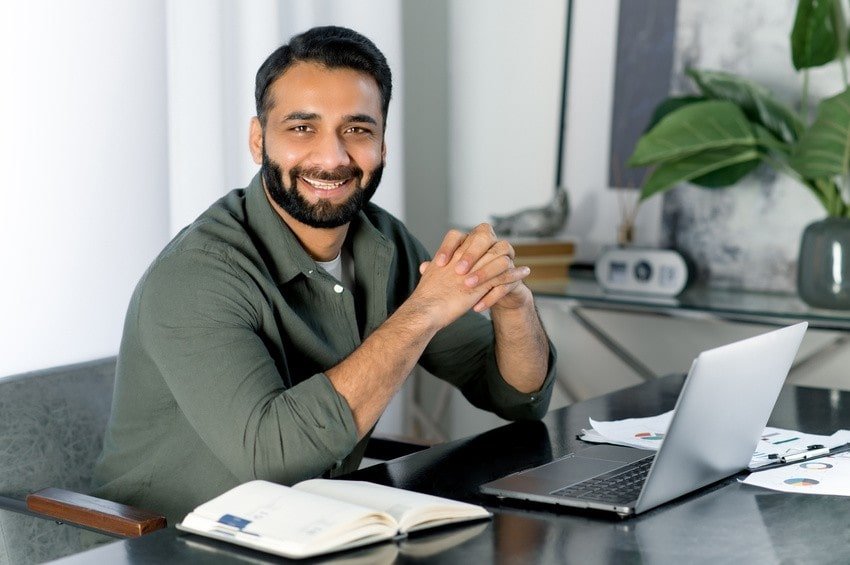 A man sits at a desk with a laptop, notebook, and papers, smiling at the camera. A plant and clock are visible in the background, suggesting he's focused on his MCCQE1 exam and with Ace QBank for his MCCQE1 exam prep.