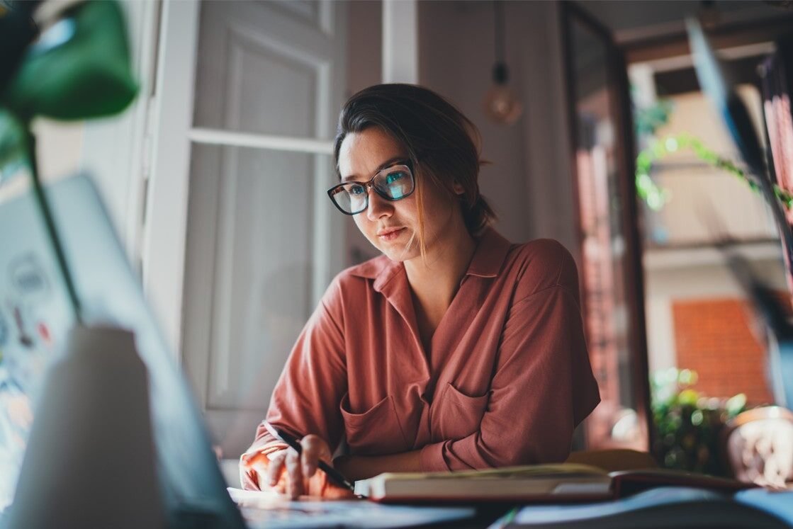 Person wearing glasses and a pink shirt sits at a desk, looking at a laptop screen, writing in a notebook with motivation, creating a study plan to study Ace QBank as natural light streams through the window.