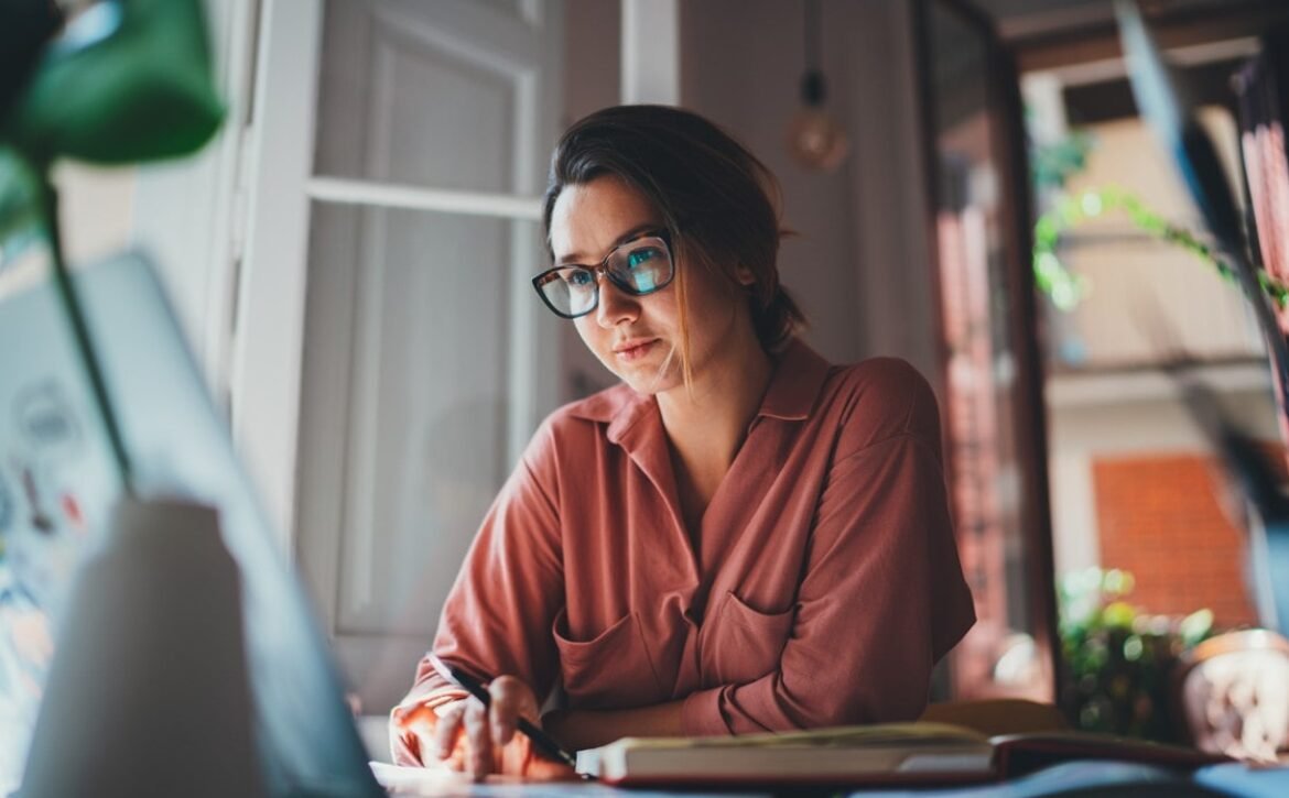 Person wearing glasses and a pink shirt sits at a desk, looking at a laptop screen, writing in a notebook with motivation, creating a study plan to study Ace QBank as natural light streams through the window.
