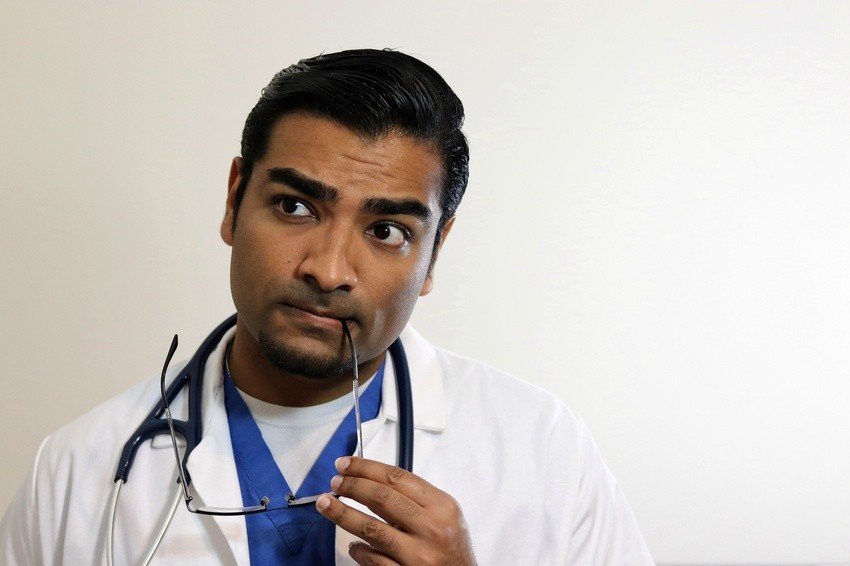 An International Medical Graduate (IMG) in a white coat with a stethoscope around his neck holds eyeglasses to his mouth and looks to the side, appearing thoughtful, perhaps considering strategies for MCCQE1 exam prep with Ace QBank.