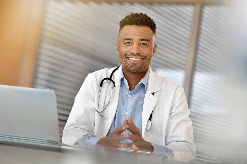 A doctor wearing a white coat and stethoscope sits at a desk with a laptop, smiling at the camera. Ready to demonstrate how to use Ace QBank effectively for MCCQE1 preparation for IMGs.