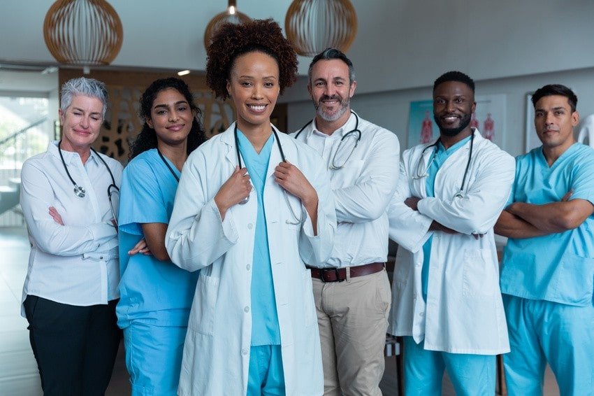 A group of six IMGs (medical professionals including doctors and nurses) stand together in a clinical setting, all wearing scrubs or white coats, have taken the MCCQE1 exam with Ace QBank and are looking confidently at the camera.
