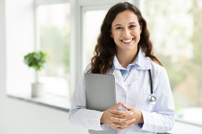 A doctor with long dark hair in a white coat, holding a clipboard and smiling, stands in a bright office with a plant in the background. Ready to share tips on MCCQE1 exam preparation.