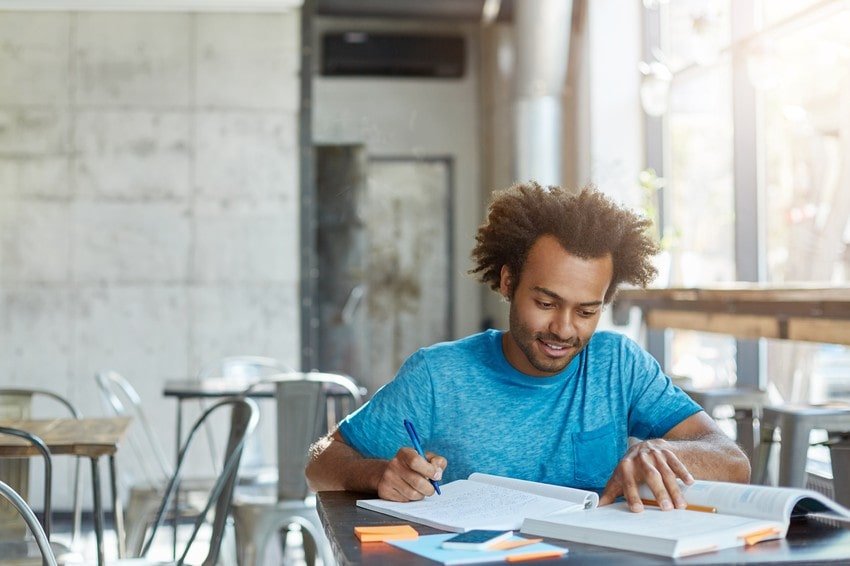 Man with curly hair wearing a blue t-shirt sits at a table in a cafe, studying for the MCCQE1 with Ace QBank, jotting notes in a notebook and reading an open book, motivation for MCCQE1 exam prep evident amid scattered sticky notes and pens.