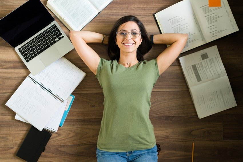 A woman lies on a wooden floor with her hands behind her head, surrounded by open notebooks. Reflecting on her MCCQE1 exam prep study plan and finding motivation to Ace QBank.